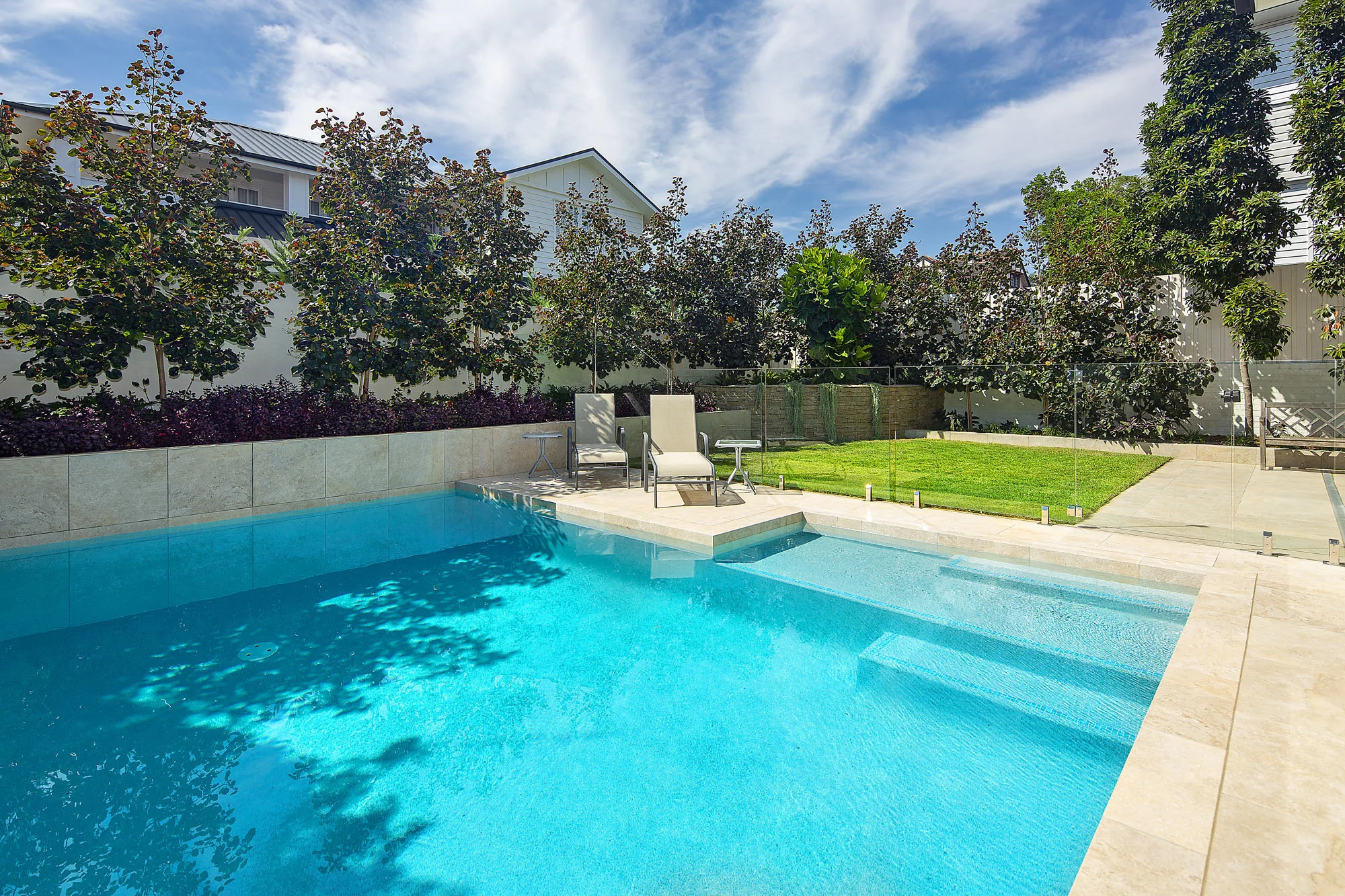 Rectangular pool with travertine coping, clipped standard trees along a rendered boundary wall, and sun loungers on the lawn