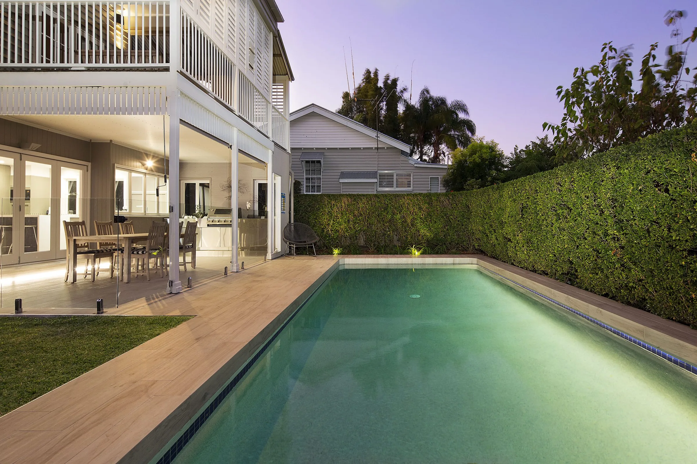 Twilight view of a lap pool and timber deck beside a weatherboard home with a dense clipped hedge wall
