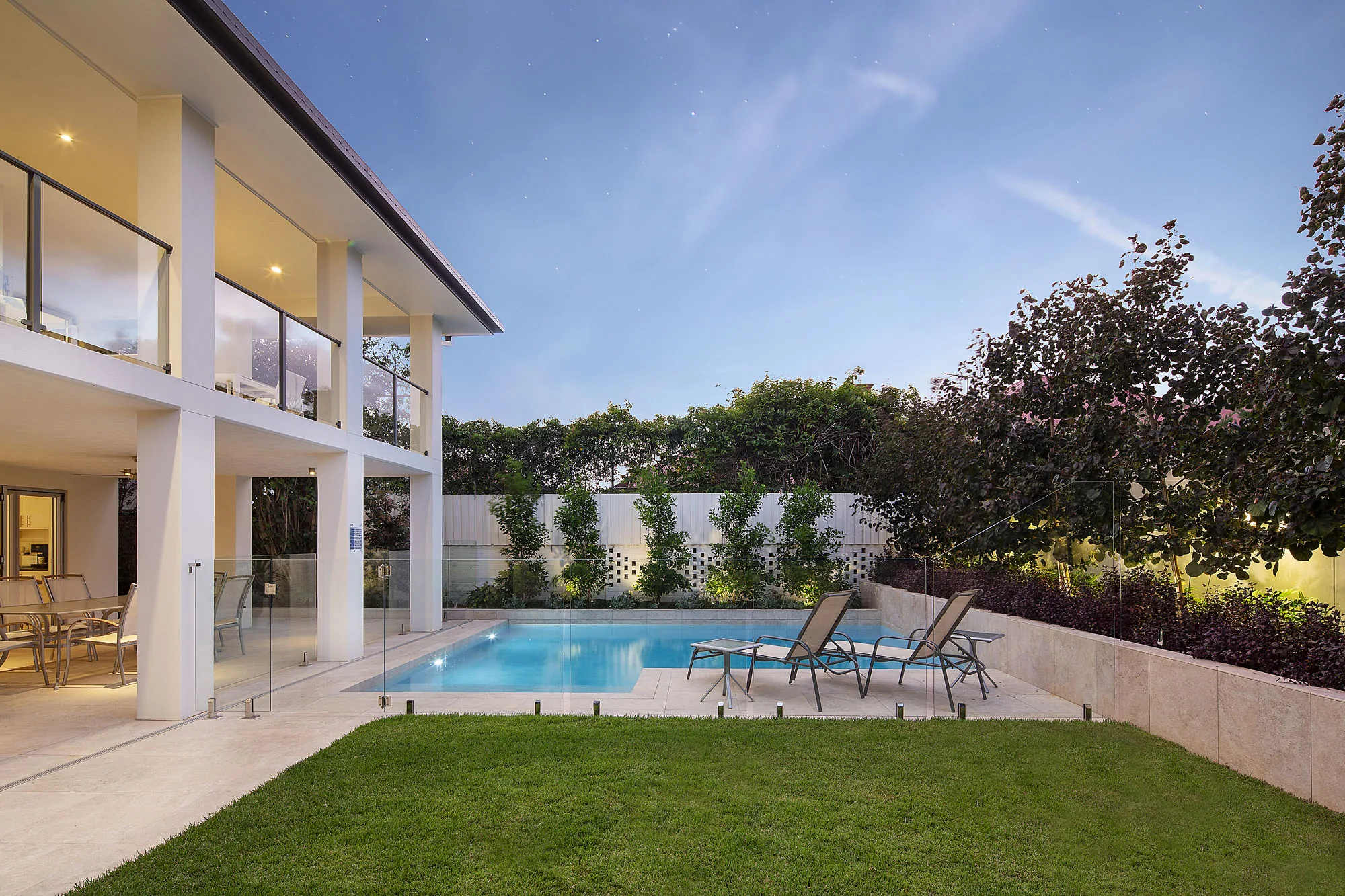 Two-storey rendered home at dusk with a rectangular pool, glass fencing, and manicured lawn