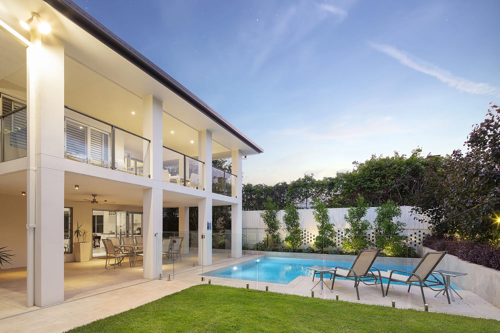 Dusk view across a rectangular pool toward a lit alfresco dining area under a rendered two-storey home