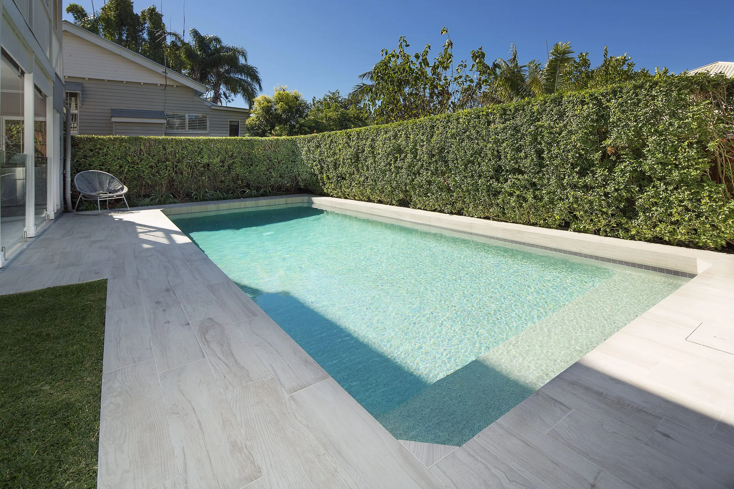 Long lap pool with a timber deck edge and a dense clipped hedge backdrop under a clear sky
