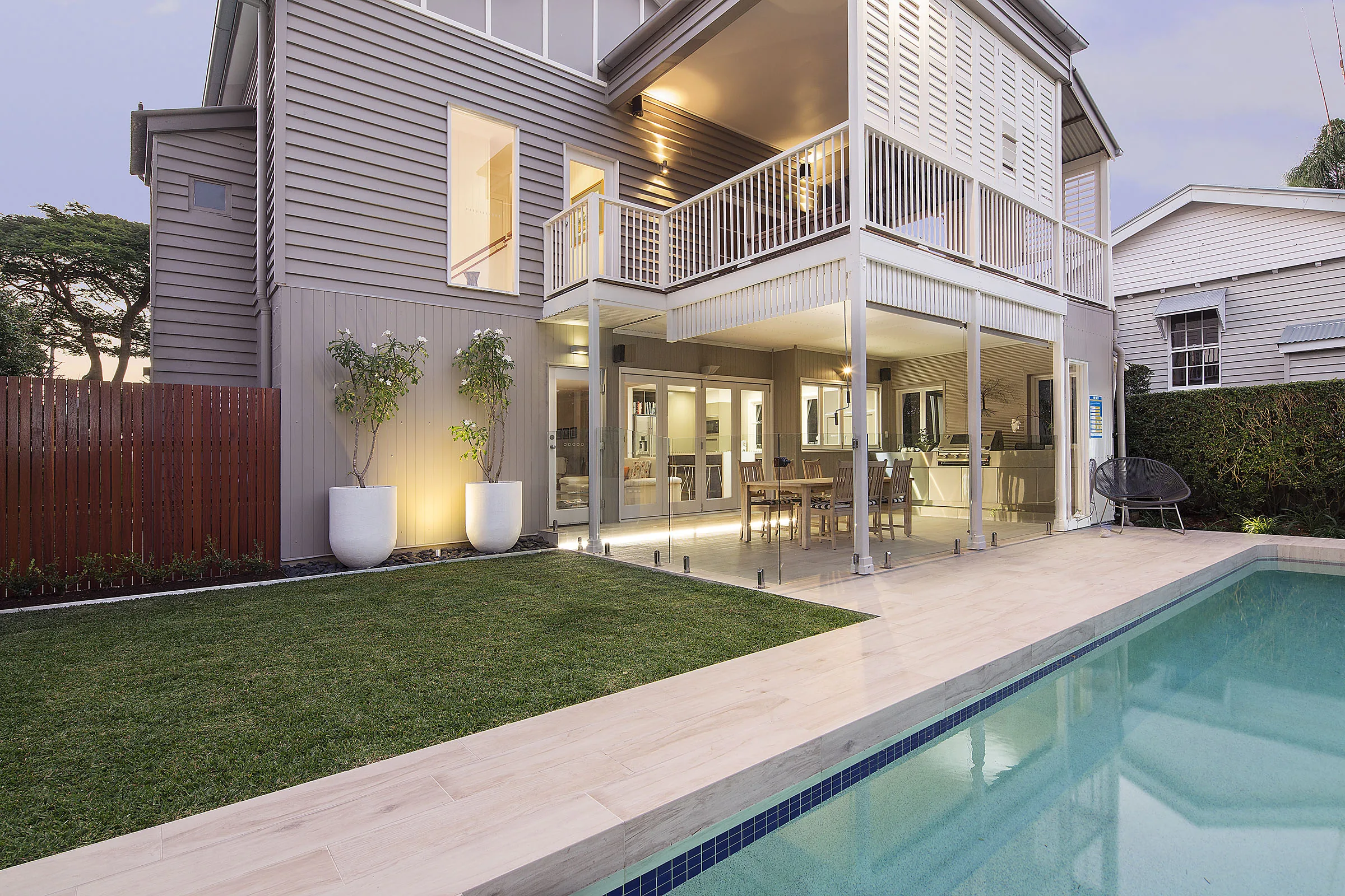Two-storey home at dusk with a rectangular pool and manicured lawn glowing under garden lighting