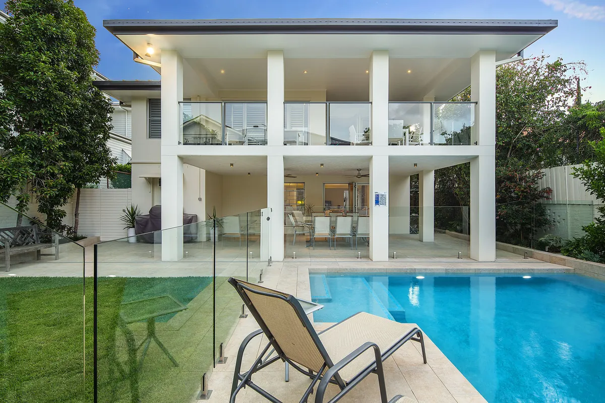 Two-storey rendered home with a cantilevered upper level, frameless glass pool fencing, and a sun lounger beside a rectangular pool