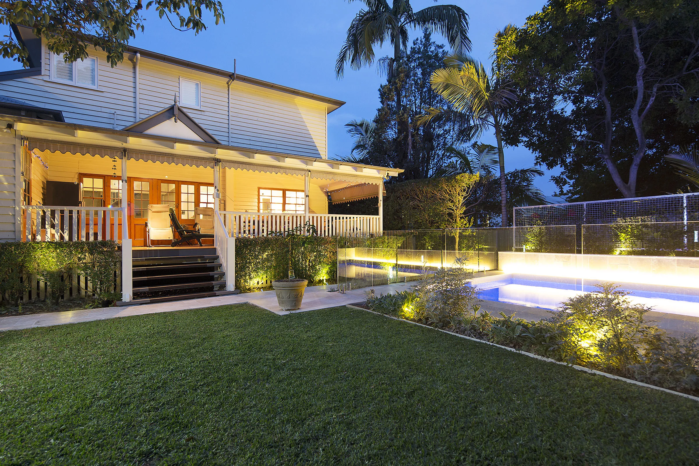 Dusk view of a rectangular pool beside a two-storey Queenslander home with a lit veranda and outdoor dining setting
