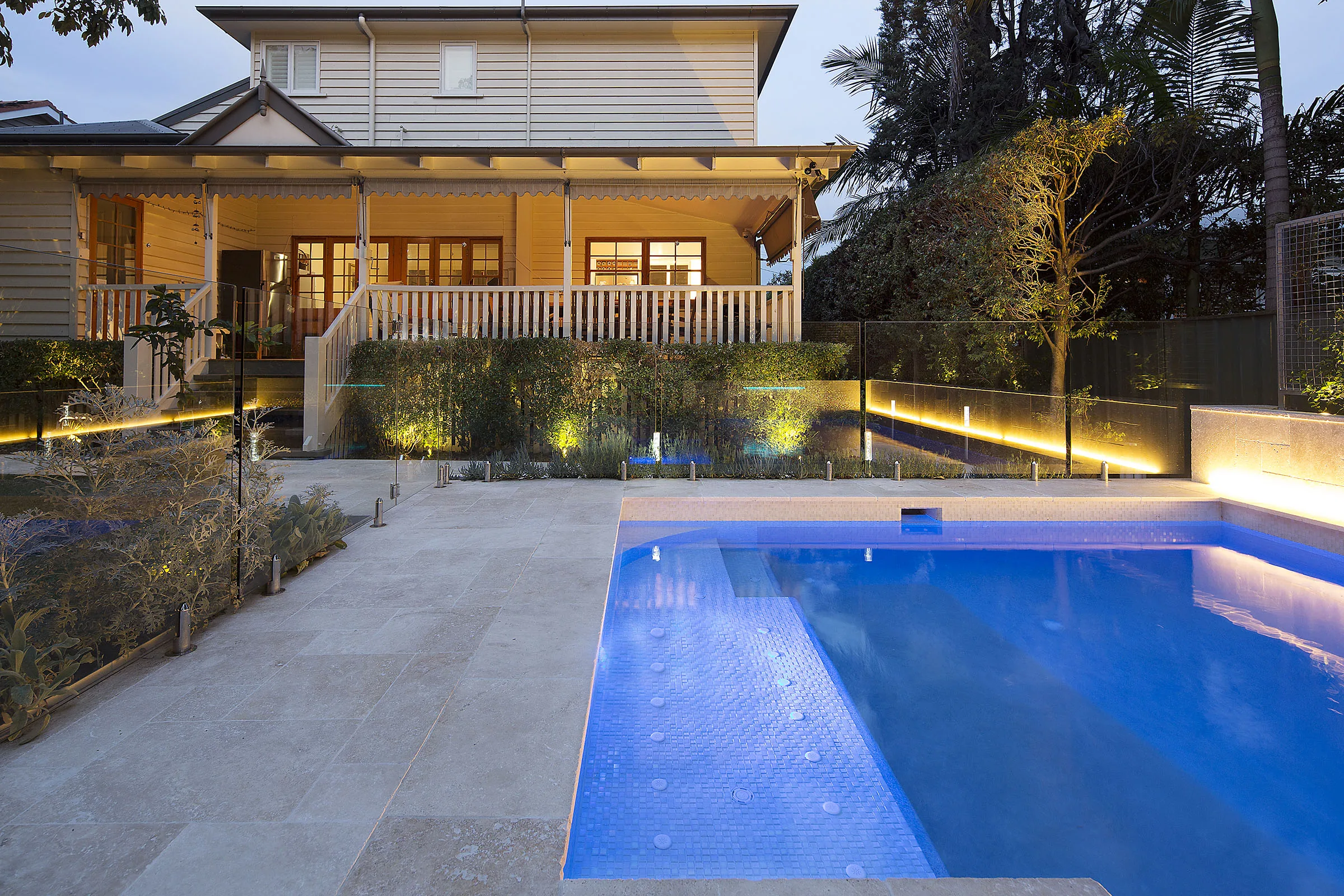 Night view of a rectangular pool with glass fencing and a two-storey Queenslander home behind