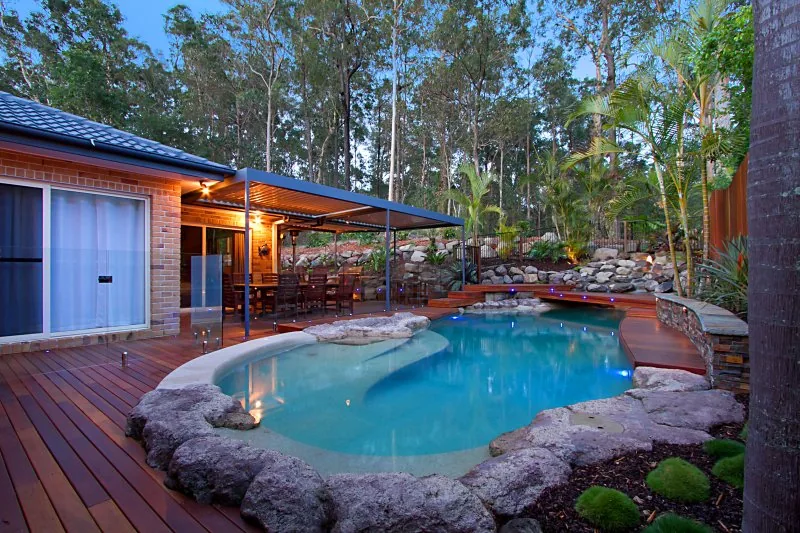 Free-form pool with boulder edging and a covered timber deck entertaining area at dusk