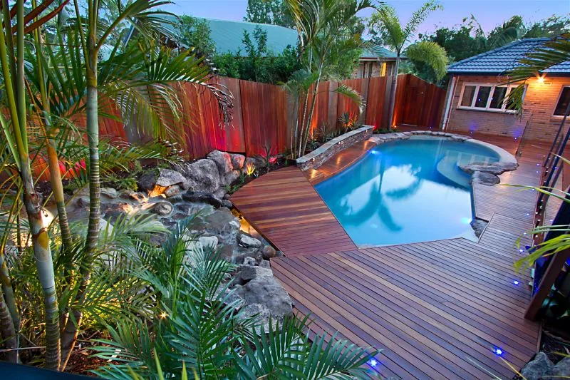 Dense golden cane and fan palms crowding a timber deck beside a free-form pool at dusk