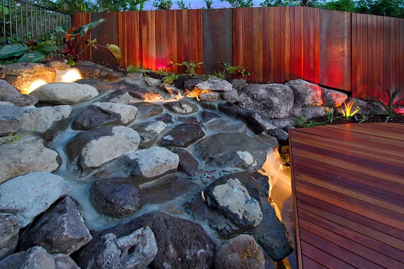 Rock waterfall feature with coloured uplighting flowing into a timber-decked pool at dusk