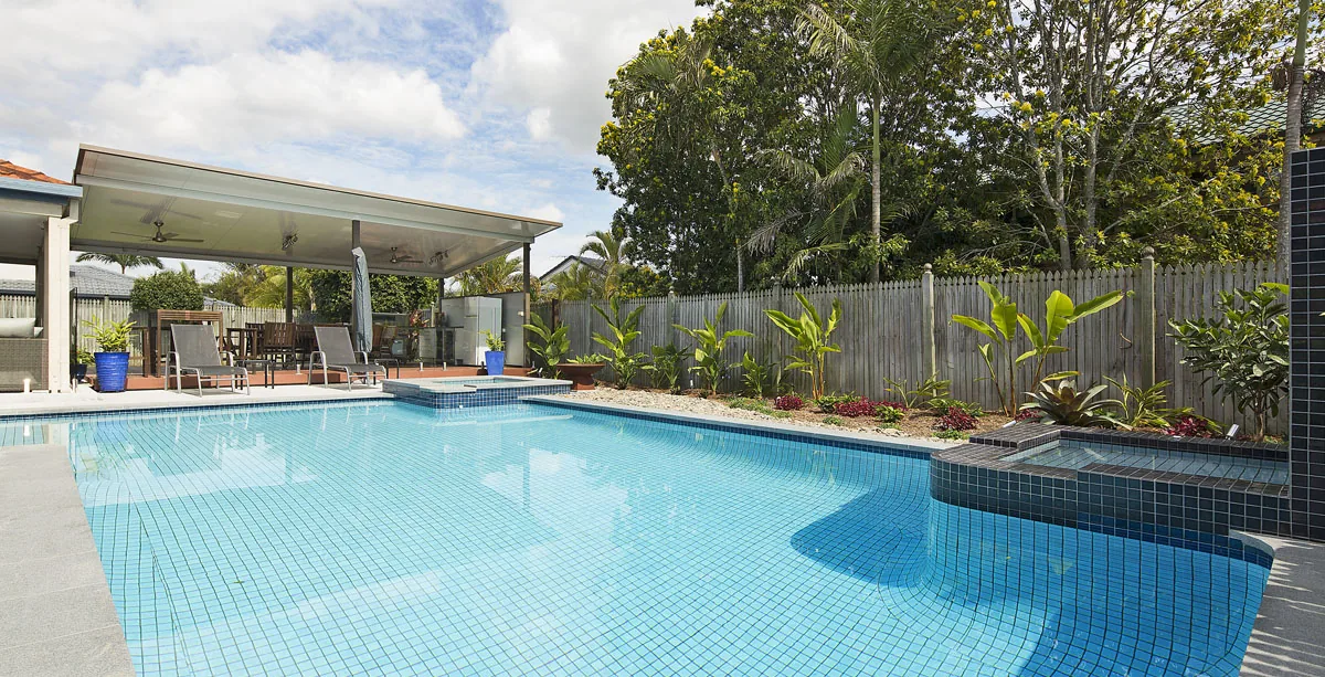 Long rectangular pool beside a covered alfresco pavilion with tropical planting along the boundary fence