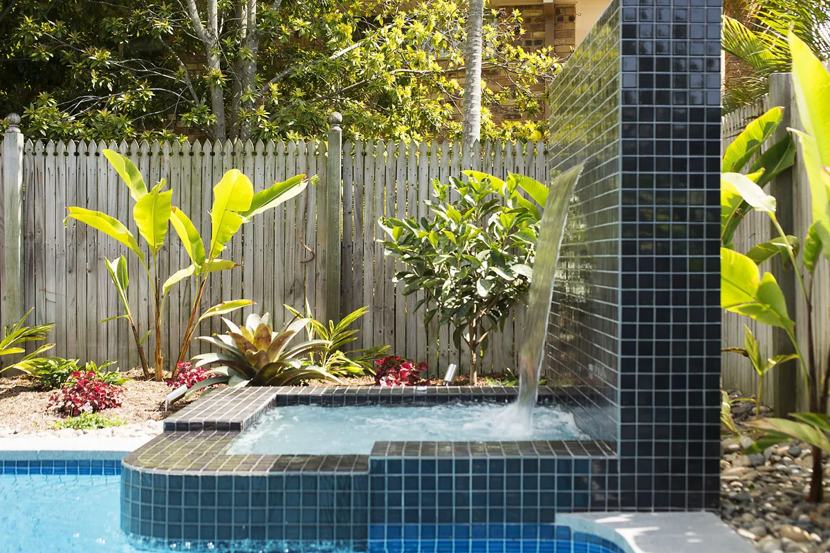 Water feature wall and sunken spa beside a pool with a lush tropical backdrop