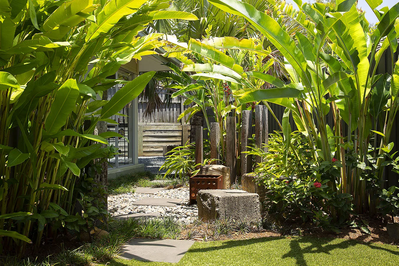 Banana leaf canopy arching over a gravel path with a corten fire crate and timber sleeper posts