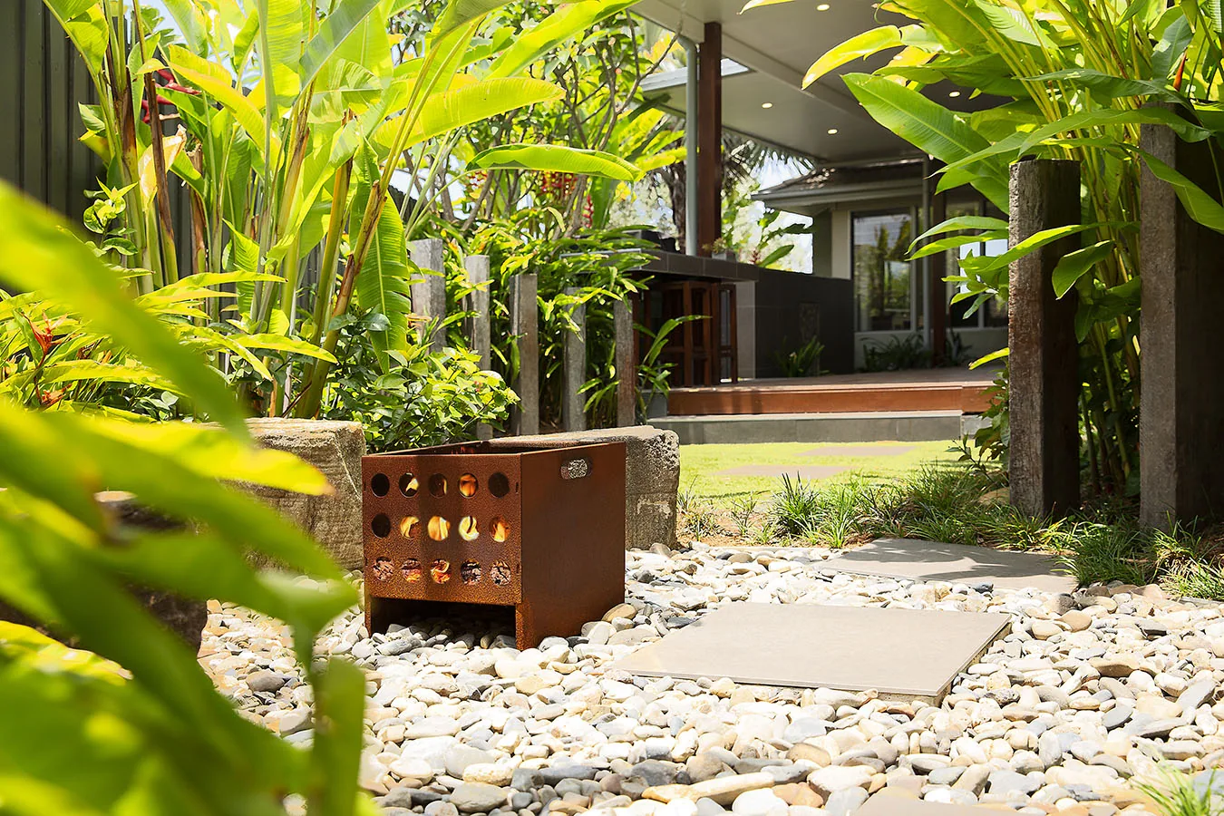 Corten fire crate glowing among dense banana leaf planting on a pebble path leading to a covered deck