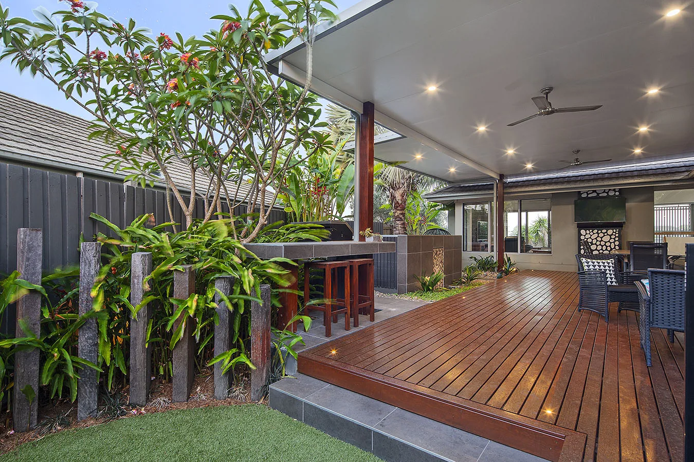 Timber deck framed by a black fretwork screen, palms, and a flowering frangipani at dusk