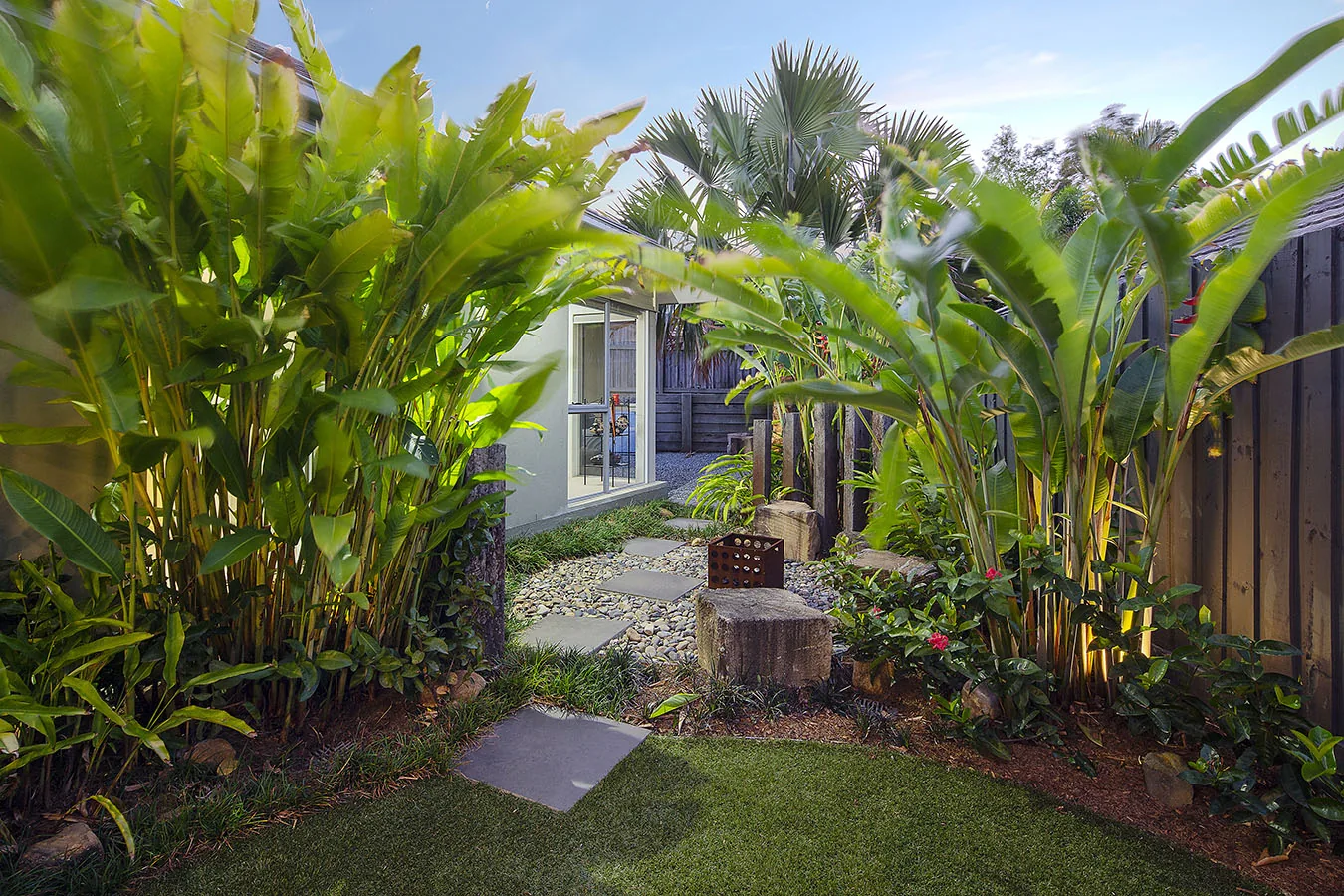 Tall bird of paradise and banana palms arching over a stepping-stone path with a corten fire crate, framing a glimpse of the home beyond